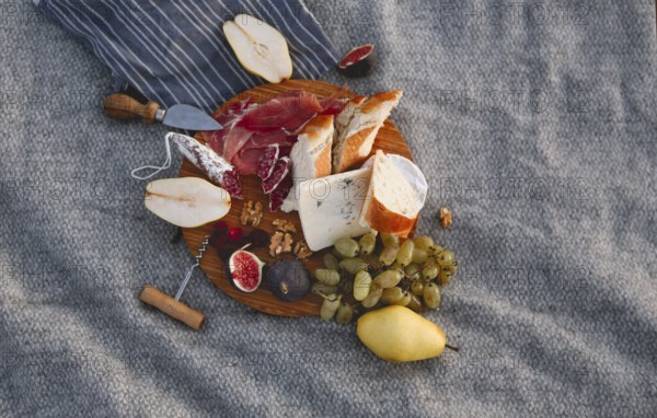 Top view of wooden board with pieces of bread pear figs nuts green grape and delicious cheese on blanket