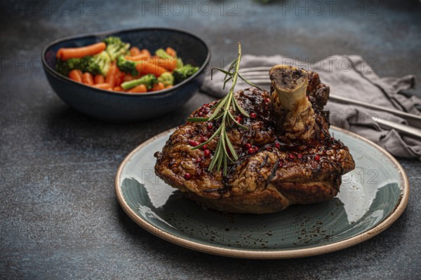 Delicious roasted ham or pork knuckle with glazed brown crispy skin, red pepper berries and rosemary on plate, steamed vegetables on rustic stone table angle view, traditional and festive meal