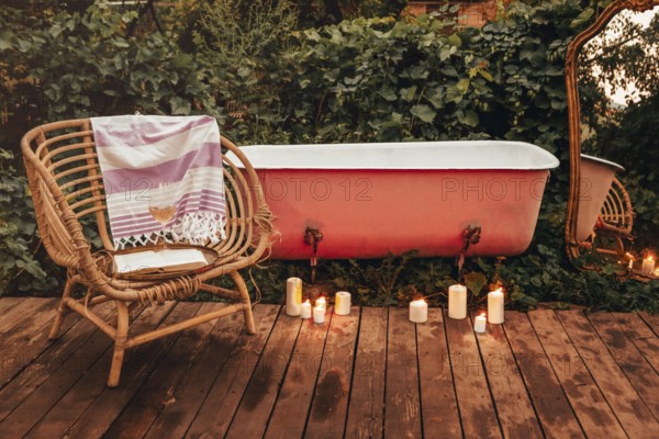 Burning candles and wicker chair with book and wine placed on wooden path near bathtub and mirror in garden