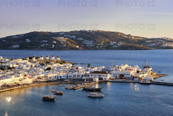Beautiful sunset view over Chora, main town of Mykonos, Cyclades, Greece and its harbor and port. Golden hour, harbor, cruises, ship, whitewashed houses and distant islands in hazy background. Vacations, leisure, Mediterranean lifestyle