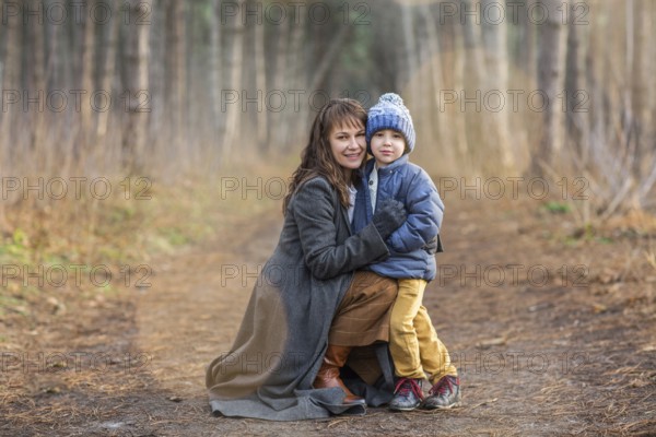 Pleased young mother hugging little son in warm wear and hat while smiling and looking at camera on empty path of autumn forest