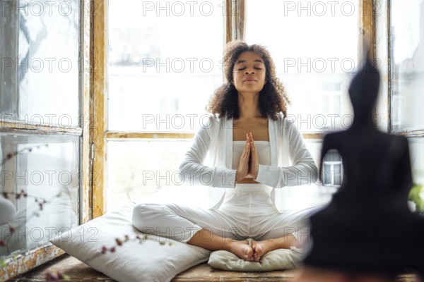Young adult happy fit slim healthy African American ethnic woman wearing white clothes sitting in yoga pose at home in living room, doing fitness morning workout exercises training. buddha silhouette