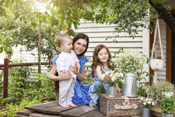 Young delighted mother sitting in armchair with adorable little siblings on terrace of country house and relaxing together during weekend