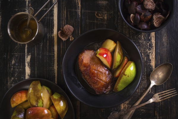 Roasted duck breast with golden crispy skin served with baked apples and chestnuts. Served on a ceramic black plate over rustic dark wooden table. Top view