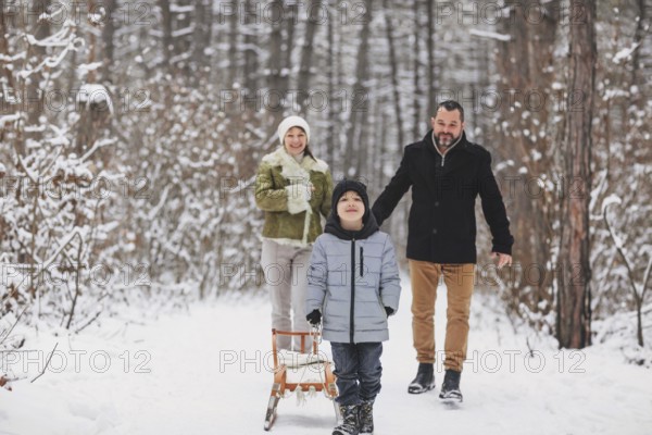 Cheerful parents walking behind glad boy with sleigh while spending time in snowy woods on winter day