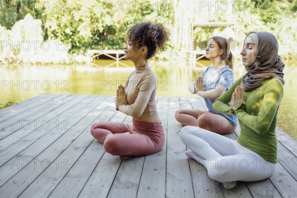 Group of teenages is engaged in fitness on pier by the lake or river. Young and positive girls go in for sports. Muslim in a hijab, african american and white females are doing stretching in the park