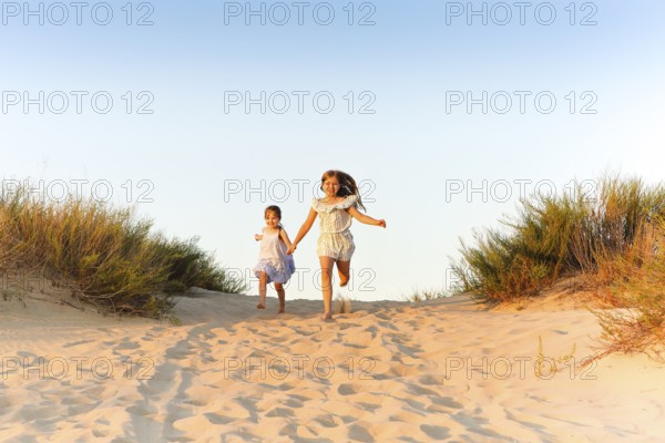 Two happy cheerful little sisters running hand in hand on sand on sunny evening beach, funny girls laughing and smiling spending time with family on seashore. Traveling and summer vacation concept