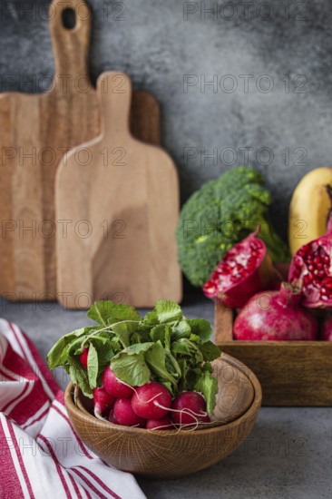 Bunch of fresh raw radish in wooden bowl on kitchen table with fresh fruit, greens, vegetables in wooden tray on grey stone background table, cooking healthy diet vegetarian food meal concept