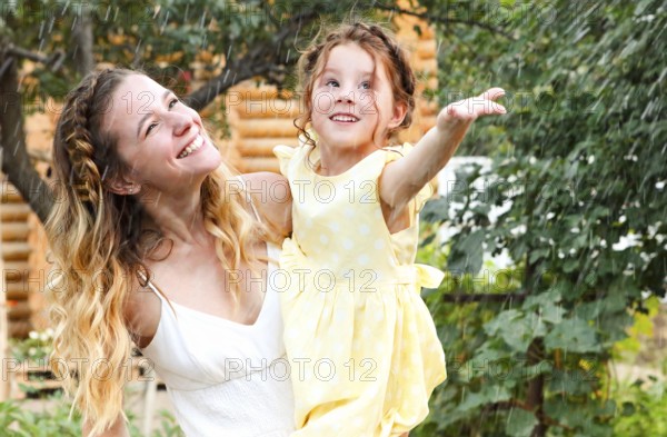 Beautiful happy family of young smiling mother and little daughter under summer rain, having fun while spending time together, mom and child playing outdoors in home garden with green trees