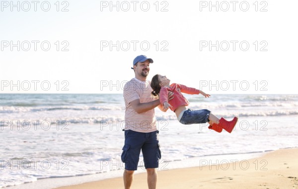 Loving father playing with his adorable little daughter on beach, swinging her around, dad and child spending time outdoors together, sunny sea in background. Parenting concept