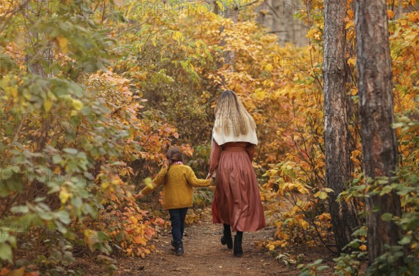 Rear view or young family, mother and little daughter, on walk in autumn forest, mom and child holding hands and walking along path covered with yellow tree leaves, spending time in beautiful nature