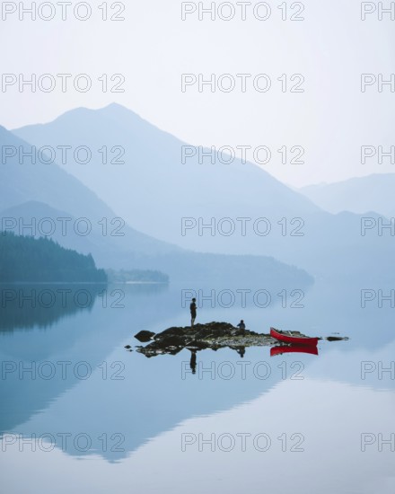 Person standing on a small island in a misty lake surrounded by mountains, with a red canoe nearby, Slocan Lake, British Columbia, Canada, North America
