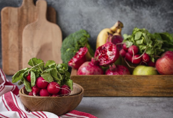 Bunch of fresh raw radish in wooden bowl on kitchen table with fresh fruit, greens, vegetables in wooden tray on grey stone background table, cooking healthy diet vegetarian food meal concept