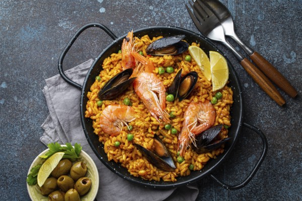 Classic dish of Spain, seafood paella in traditional pan on rustic blue concrete background top view. Spanish paella with shrimps, clamps, mussels, green peas and fresh lemon wedges from above