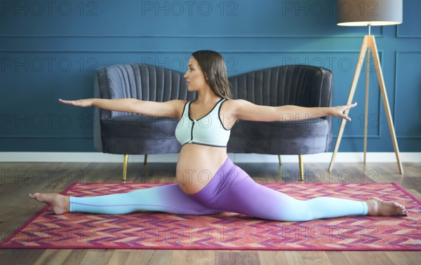 Pregnant sporty woman practicing yoga at home doing stretching exercise the side split pose Hanumanasana. Conscious motherhood. Healthy lifestyle and baby care concept