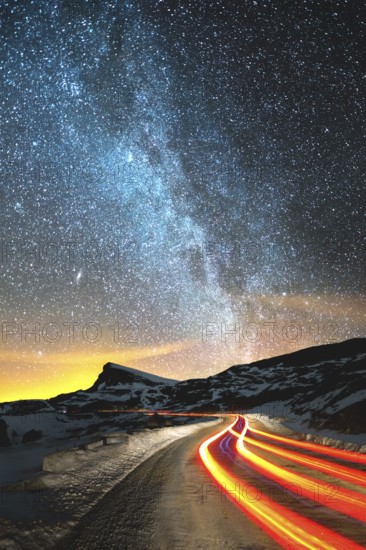 Night landscape. Night sky with a north hemisphere Milky Way and stars. The night road illuminated by the car winds with a serpentine and leaves in a distance to a foot of an acute rock. Light trails of red color. The concept of the path to knowledge and