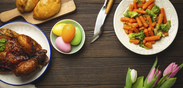 Easter food on dark rustic table: pastel colored eggs, roasted chicken and vegetables, buns and spring flowers tulips top view flay lay, Easter family dinner meal with festive dishes, space for text
