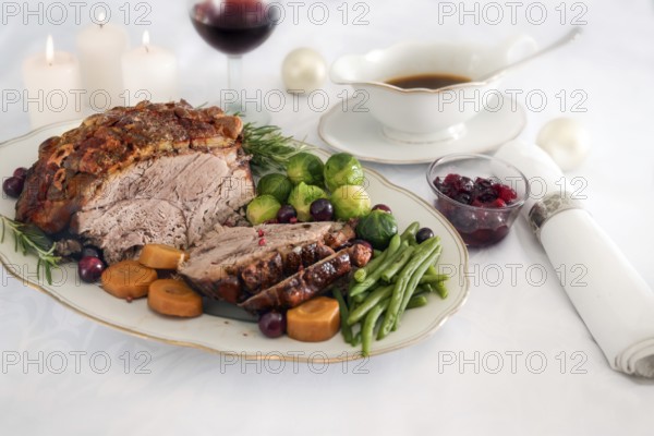 Roast pork ham with crust, green beans, Brussel sprouts, carrots, cranberry and sauce on white table cloth with candles and Christmas decoration, festive dinner, selected focus, narrow depth of field