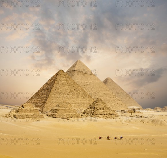Dramatic sunset behind all distant Egyptian Pyramids together with line of camels walking in foreground in Giza, Cairo, Egypt. Wide compressed telephoto shot with plenty of copy space