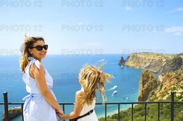 Mother and little girl standing next to fence on viewpoint and admiring picturesque view of rocky sea coast in sunny day with clear sky and blue water