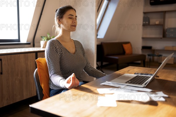 Stress relief during freelance work. Brunette woman freelancer sitting in lotus pose and meditating with hands in mudra gesture on chair in front of open laptop on table, working from home