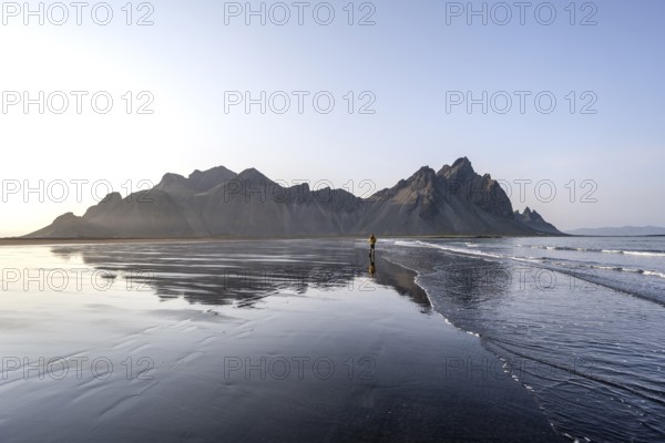 Tourist at black lava beach, sandy beach and sea, mountains Klifatindur reflected in the water, Eystrahorn and Kambhorn, headland Stokksnes, mountain range Klifatindur, Austurland, East Iceland, Iceland, Europe