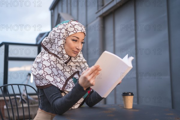 Arab girl with white veil at the computer on the terrace of a coffe shop, having a lot of fun reading a beautiful book