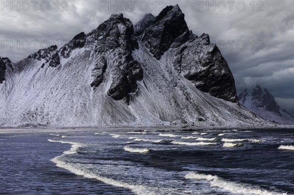 White foamy waves on the beach of Stokkness, behind snowy rocky slopes of Vestrahorn, near Höfn, Sudausturland, Iceland, Europe