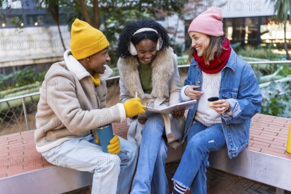 Three cheerful young students are sitting on a bench, studying and having a coffee break outdoors in the city during wintertime