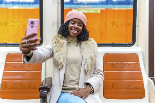 Young woman dressed in winter attire, smiling and taking a selfie while enjoying a ride on a subway train in the city