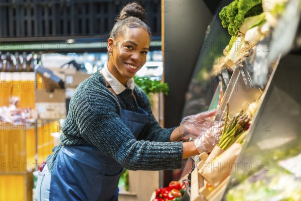 African american saleswoman wearing plastic gloves arranging organic asparagus at the vegetable counter of a supermarket
