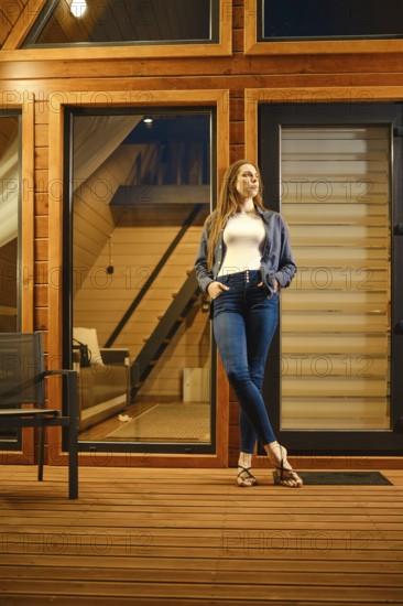 A beautiful brunette woman stands on the doorway of a large wooden cabin in the evening