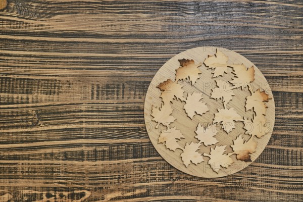A circular wooden base hot pad displays intricately cut wooden leaves with varying hues, arranged in an artistic pattern