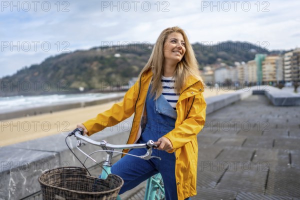 Young woman wearing yellow raincoat riding bicycle on boardwalk by the beach, enjoying ocean view and smiling