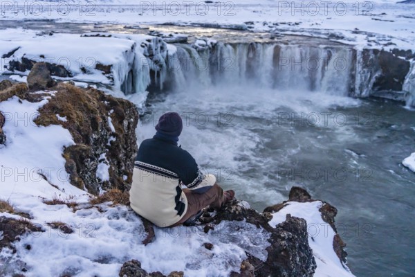 A man hiker sitting looking at the frozen Godafoss waterfall at sunset in winter, Iceland, Europe
