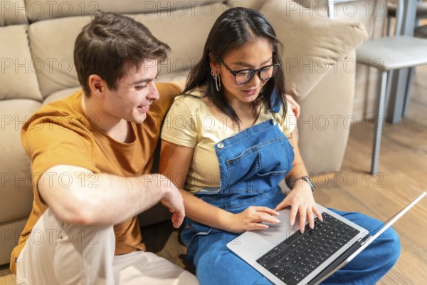 Young multiethnic couple sitting on floor and using laptop, browsing internet and smiling in cozy living room