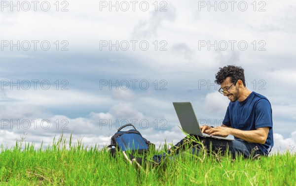 Smiling man with laptop sitting on the grass in the field. Side view of man sitting on green grass working with his laptop. Smiling freelancer guy working from the field