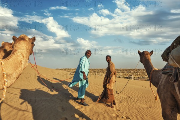 Rajasthan travel background, two indian cameleers (camel drivers) with camels in dunes of Thar desert. Jaisalmer, Rajasthan, India, Asia
