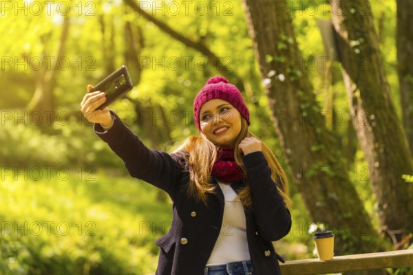 Young man with a black jacket, scarf and red woolen hat enjoying in an autumn park, taking a selfie with the mobile, at sunset