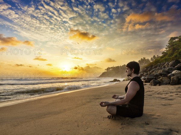 Young sporty fit man doing yoga meditating in padmasana lotus pose on tropical beach on sunset