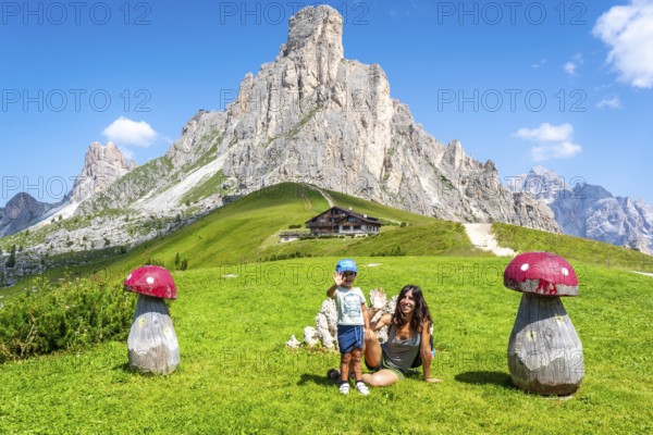 Mother and son waving near big wooden mushrooms with scenic view of nuvolau peak and mountain hut at giau pass in dolomites