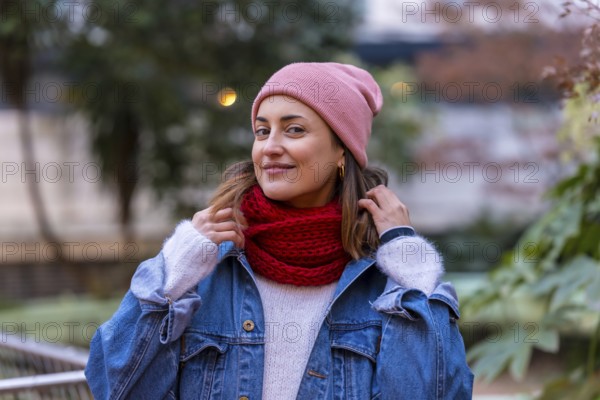 Happy woman wearing stylish winter clothes, playfully adjusting her red scarf while enjoying a stroll through a serene city park