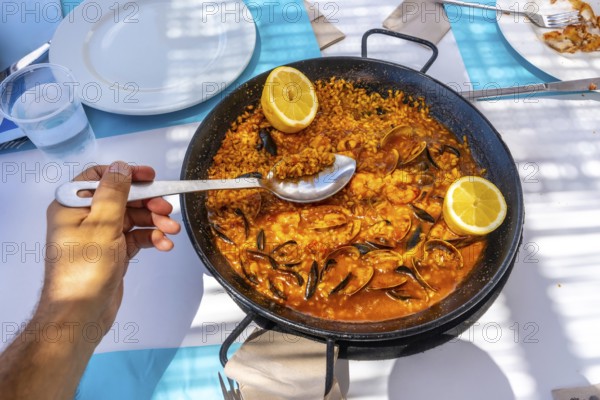 Hand holding spoon scooping seafood paella from pan in outdoor restaurant during summer vacation