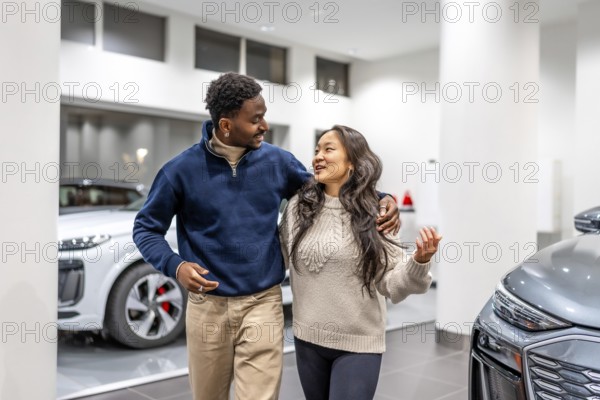Happy multiethnic couple walking and choosing an electric car in a dealership, hugging and smiling