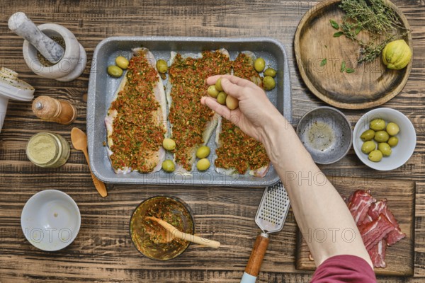 A hand adds green olives to a tray of seasoned fish fillets covered in vibrant herbs and spices. Surrounding the dish are various ingredients, creating a warm cooking atmosphere