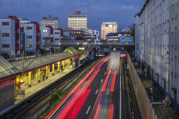 Essen, North Rhine-Westphalia, Germany, motorway A40 in the city centre, exit Essen Zentrum, during dusk. People waiting at Savignystrasse underground station. On the right, residential buildings behind a noise barrier. There is a LANUV air monitoring station, Essen Kruppstrasse 117 (EKRU2), on the right under the lantern. Residents are affected by noise and exhaust fumes. Building with Postbank, Eon and Evonik lettering at the back, Europe