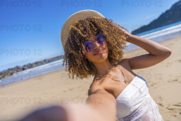 Personal perspective of a cute beauty latin woman in summer clothes, sunglasses and sun hat taking selfie on the beach