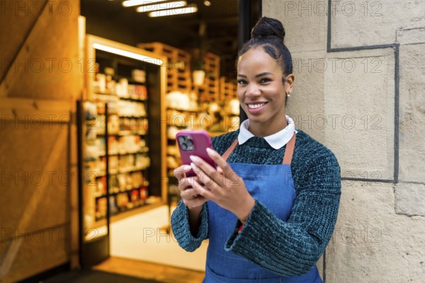 Happy african american saleswoman wearing apron using smartphone at organic supermarket entrance