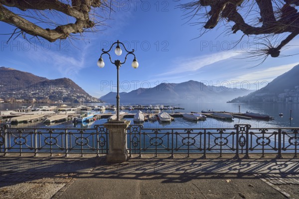 Lantern, forged railing, lakeside promenade, shadow, marina, trees, motorboats, hill with village, haze, backlight, blue sky with veil clouds, Lake Lugano, Lugano, district Lugano, canton Ticino, Switzerland, Europe