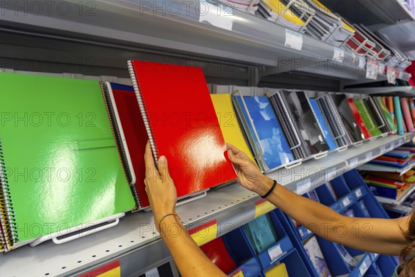 A person is holding a green spiral notebook in a store. There are many notebooks on the shelves, including some that are red and yellow. Back to school concept